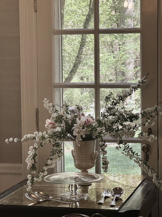 graceful arching white flowers and white and red tulips in vase in elegant setting near a window with woods